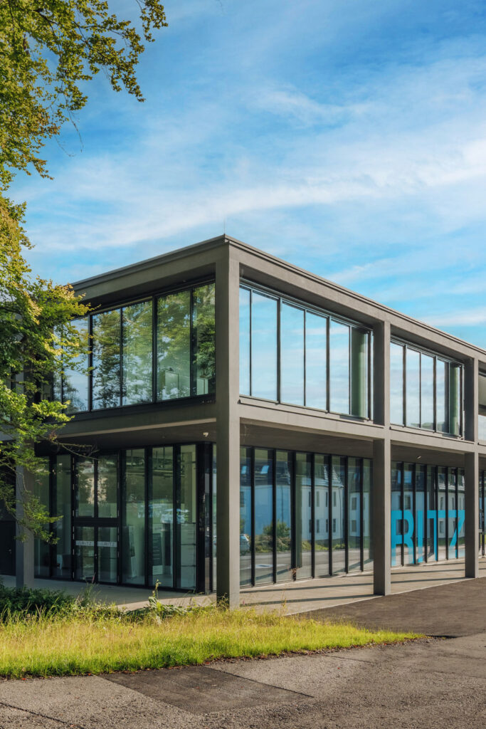 Eingangsbereich des RITZ Innovationszentrums mit moderner Glasfassade, davor grüne Wiese und geteerte Straße.