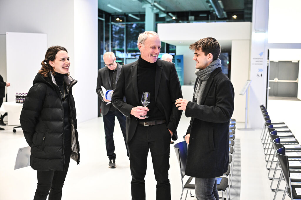 Drei Personen im Gespräch bei der Veranstaltung "Beispielhaftes Bauen", eine Person hält ein Glas in der Hand, im Hintergrund weitere Gäste in modernem, hell beleuchtetem Raum.