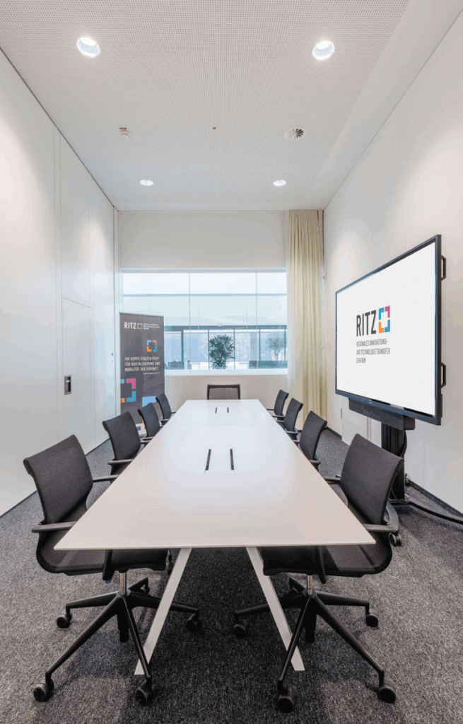Moderner Besprechungsraum mit großem Konferenztisch, neun Bürostühlen, Bildschirm mit RITZ-Logo und Roll-Up, grauem Teppichboden und Tageslicht durch Innenfenster.
