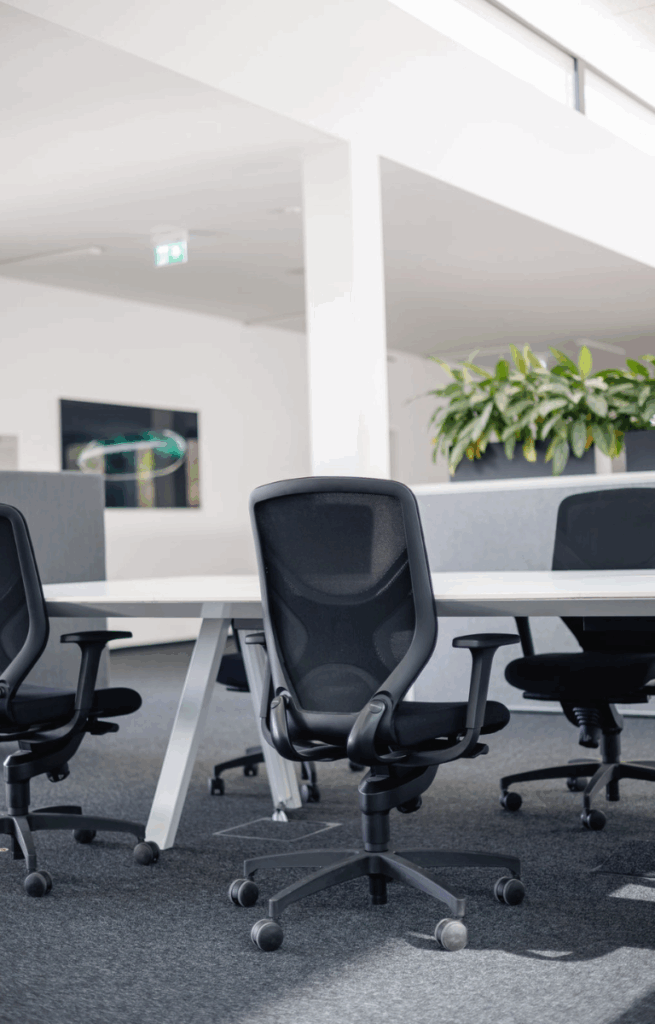 Nahaufnahme eines ergonomischen Bürostuhls vor einem weißen Schreibtisch in einem offenen, hellen Coworking-Bereich mit Pflanzen im Hintergrund.