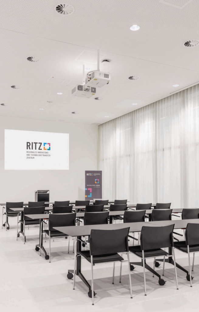Seminarraum mit Tischreihen und Stühlen in Frontalbestuhlung, Projektor zeigt RITZ-Logo auf der Leinwand.