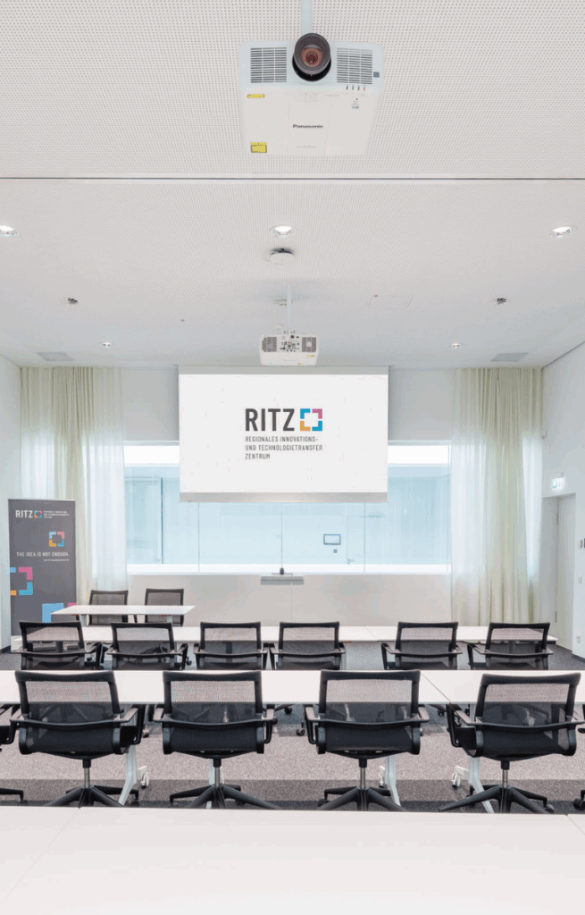 Moderner Meetingraum mit Leinwand, Projektor und mehreren Bürostühlen in Reihenanordnung, Blickrichtung zur Präsentationsfläche.