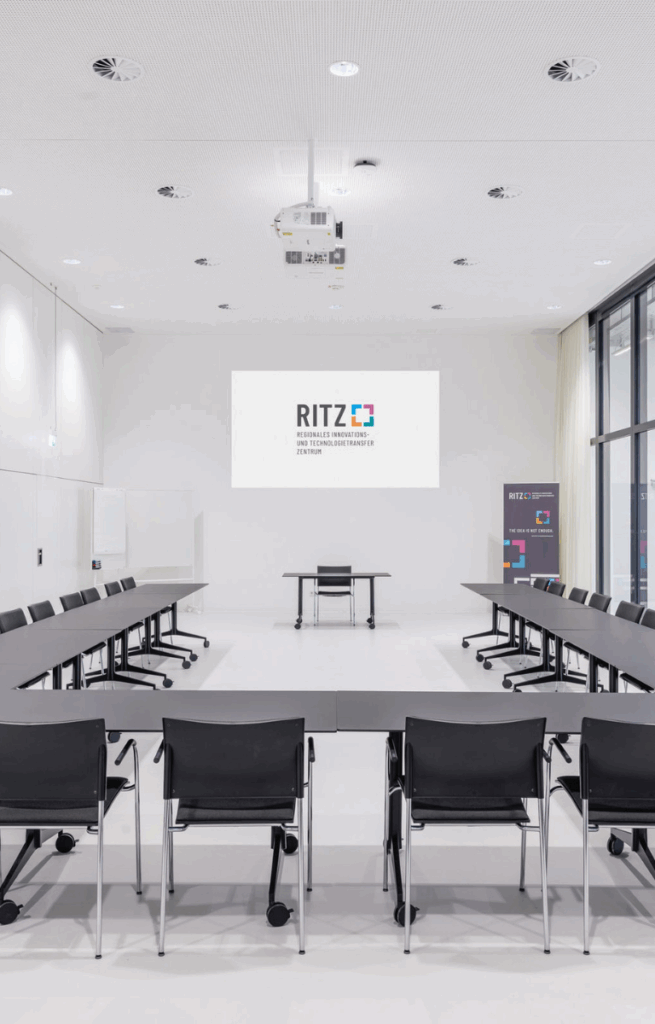 Eventraum mit Präsentationsfläche und Bestuhlung in U-Form, moderner Deckenbeamer und große Projektionsfläche mit RITZ-Logo.