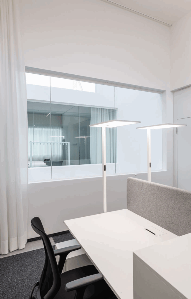 Zwei moderne Schreibtischarbeitsplätze mit LED-Stehlampen vor einer Glaswand im hellen Büroraum.