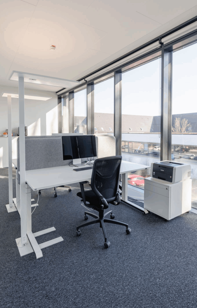 Heller Einzelarbeitsplatz mit Bildschirm, ergonomischem Stuhl und Blick durch bodentiefe Fenster auf ein Nachbargebäude.