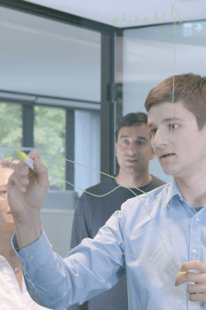 Mann in hellblauem Hemd skizziert ein Diagramm mit gelbem Marker auf eine Glasscheibe, während zwei weitere Personen aufmerksam zusehen.