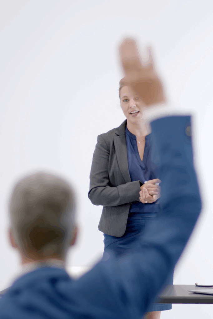 Frau im Business-Outfit hält einen Vortrag, während eine Person im Vordergrund die Hand hebt, um eine Frage zu stellen.