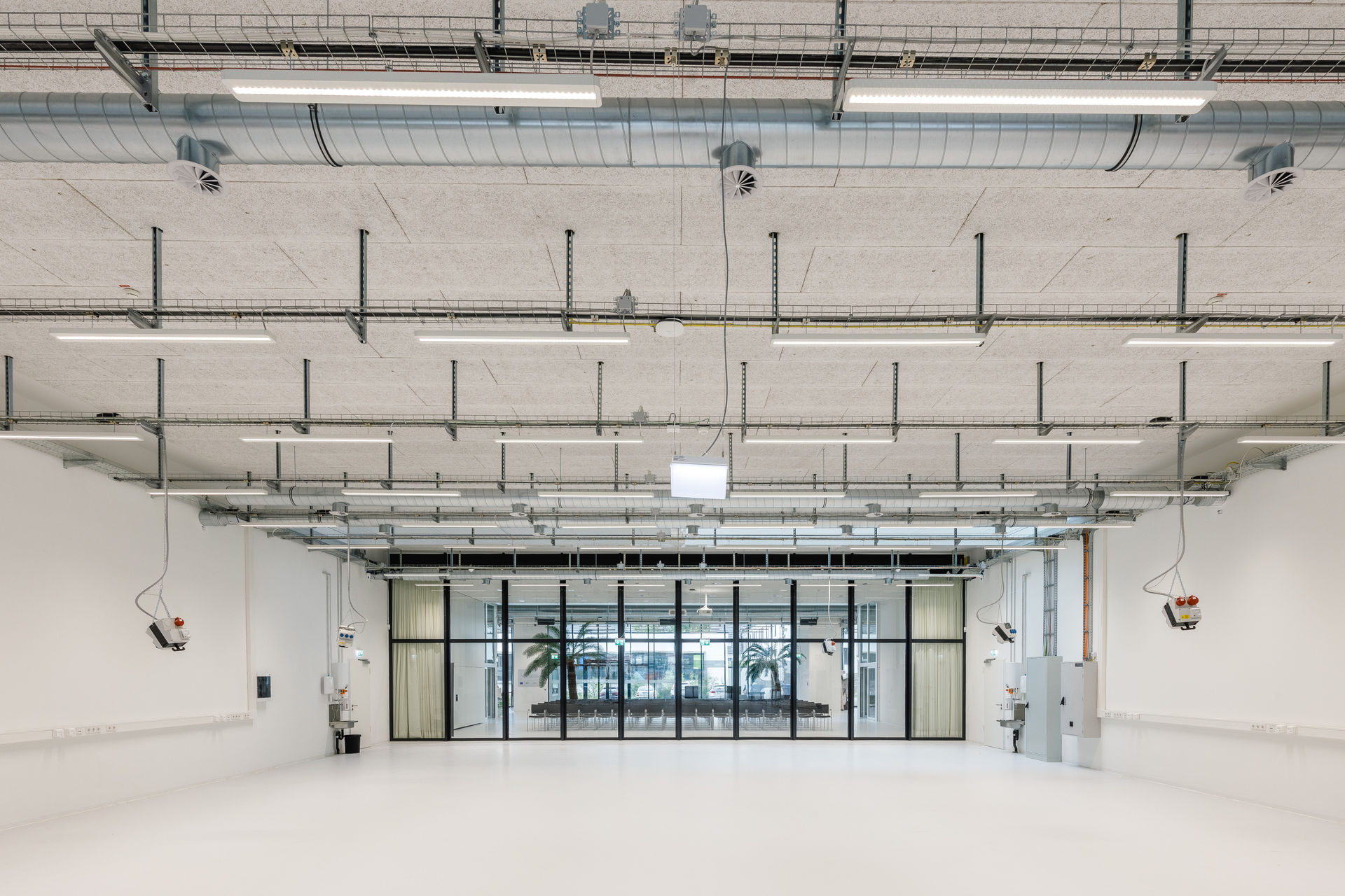 Blick auf einen großen, leeren Technikraum mit weißem Boden und weißen Wänden. An der Decke verlaufen offen installierte Kabeltrassen, Lüftungsrohre und LED-Leuchten. Mehrere Strom- und Druckluftanschlüsse hängen von der Decke. Die Rückwand besteht aus einer großflächigen Glasfront mit schwarzem Rahmen und Blick in einen angrenzenden Raum mit Palmen und Bestuhlung. Entlang der Seitenwände sind Steckdosenleisten und technische Installationen angebracht.
