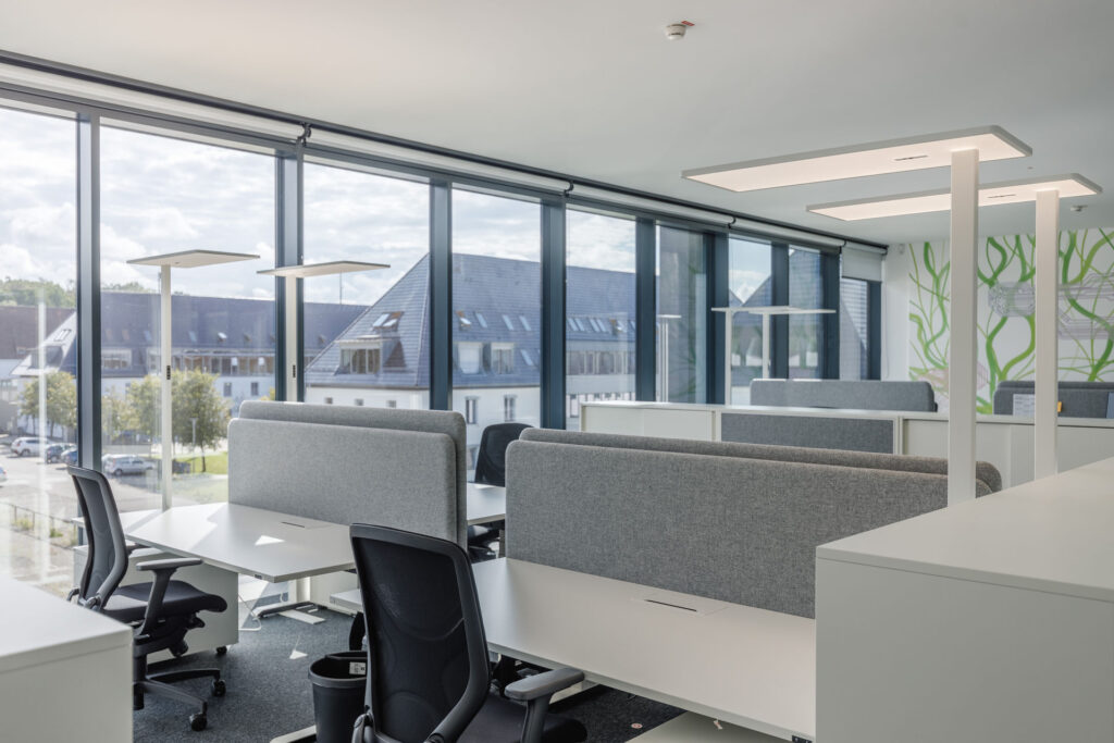 Heller, moderner Open Space im RITZ mit höhenverstellbaren Schreibtischen, grauen Akustik-Trennwänden, ergonomischen Bürostühlen, großen Fensterfronten und einer Wandgestaltung mit grün geschwungenen Linien, die an stilisierte Pflanzenranken und Schaltkreise erinnert.