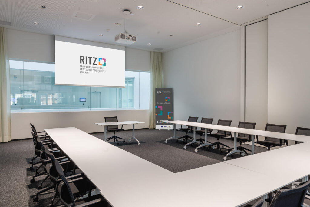Moderner Konferenzraum im RITZ mit U-förmiger Tischanordnung, Bürostühlen, Beamer und Leinwand mit RITZ-Logo sowie einem Roll-Up-Banner in der Ecke.