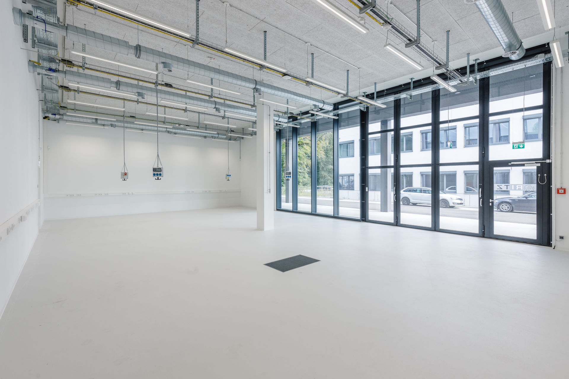 Großer, leerer Technikraum mit weißem Boden und weißen Wänden. An der Decke verlaufen sichtbare Kabeltrassen, Lüftungsrohre und LED-Leuchten. Mehrere Versorgungsanschlüsse hängen von der Decke herab. Entlang der linken Wand verläuft eine Reihe von Steckdosenleisten. Auf der rechten Seite befindet sich eine breite Fensterfront mit Glastür, die den Blick nach draußen auf parkende Autos und das gegenüberliegende Gebäude freigibt. Ein einzelner Stützpfeiler steht frei im Raum.