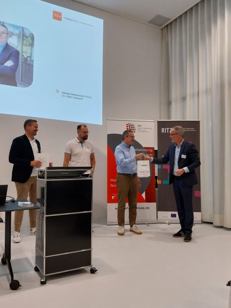 Dankesmoment auf der Bühne beim Production Excellence Day 2024 im RITZ. Zwei Männer schütteln sich die Hand, flankiert von Roll-ups der Partnerorganisationen.