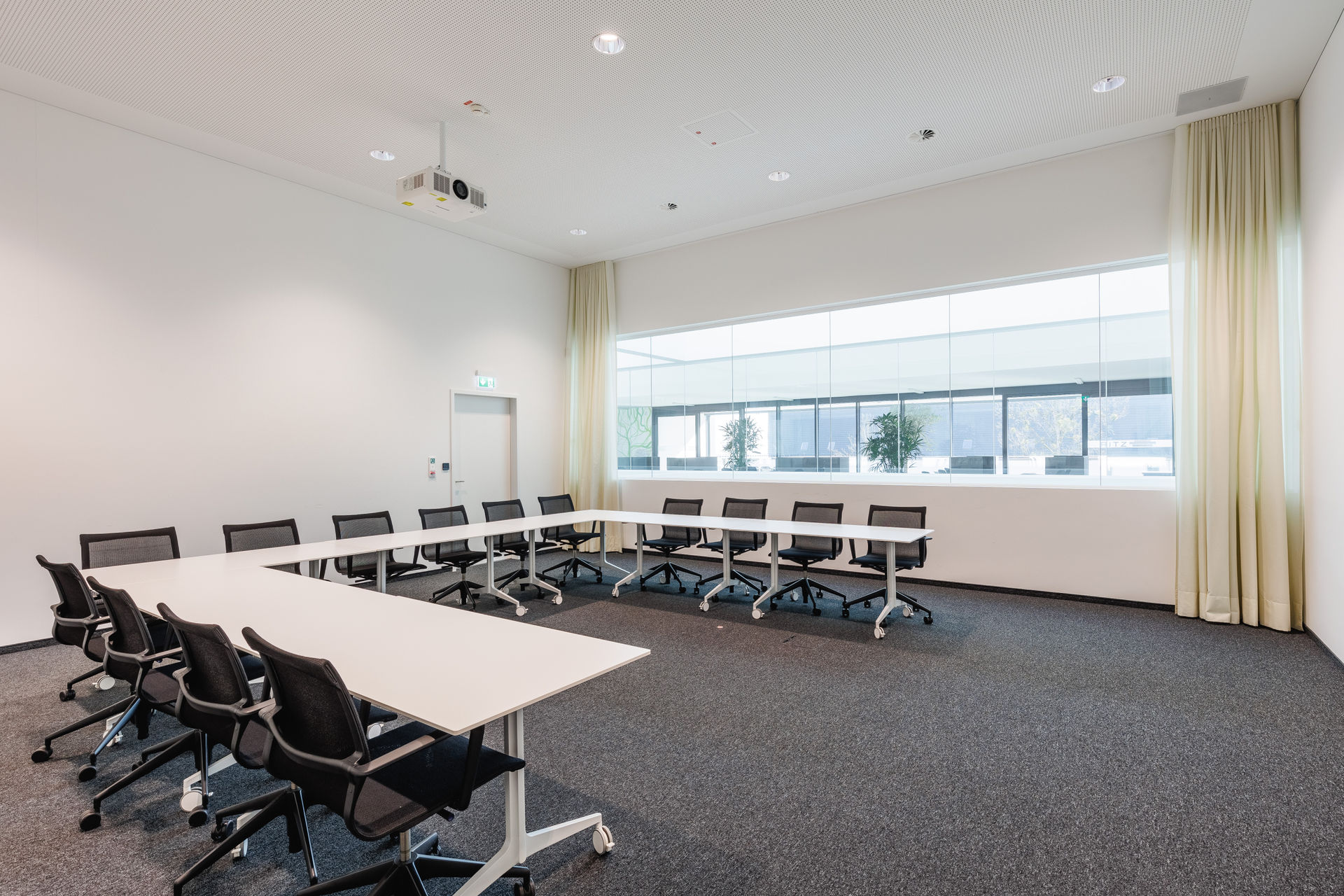 Zu sehen ist ein rechteckiger, heller Besprechungsraum mit Teppichboden und U-förmig angeordneten Konferenztischen auf Rollen. Die schwarzen Bürostühle sind ergonomisch geformt und ebenfalls mobil. An der Decke ist ein Beamer montiert, an der Wand eine Tür mit Fluchtwegbeschilderung. Die rechte Wandseite wird durch ein breites, raumhohes Fenster durchbrochen, das in einen angrenzenden Raum mit Pflanzen und Tageslichtblick führt. Links und rechts vom Fenster hängen helle Vorhänge. Die Decke ist akustisch wirksam perforiert.