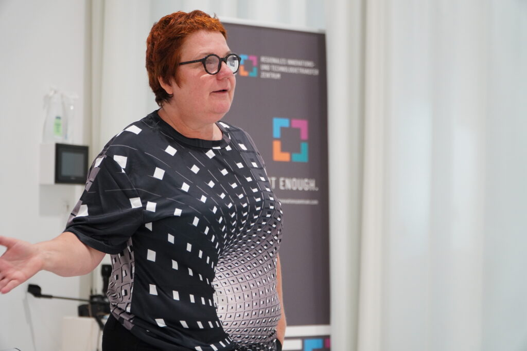 Anne-Claire Schroeder-Rose steht gestikulierend vor einem Roll-up des RITZ Innovationszentrums und hält einen Vortrag. Sie trägt ein schwarzes Shirt mit geometrischem Muster. Die Szene stammt aus dem Workshop „Innovationsgeist schärfen durch die Methodik von Better by art“.
