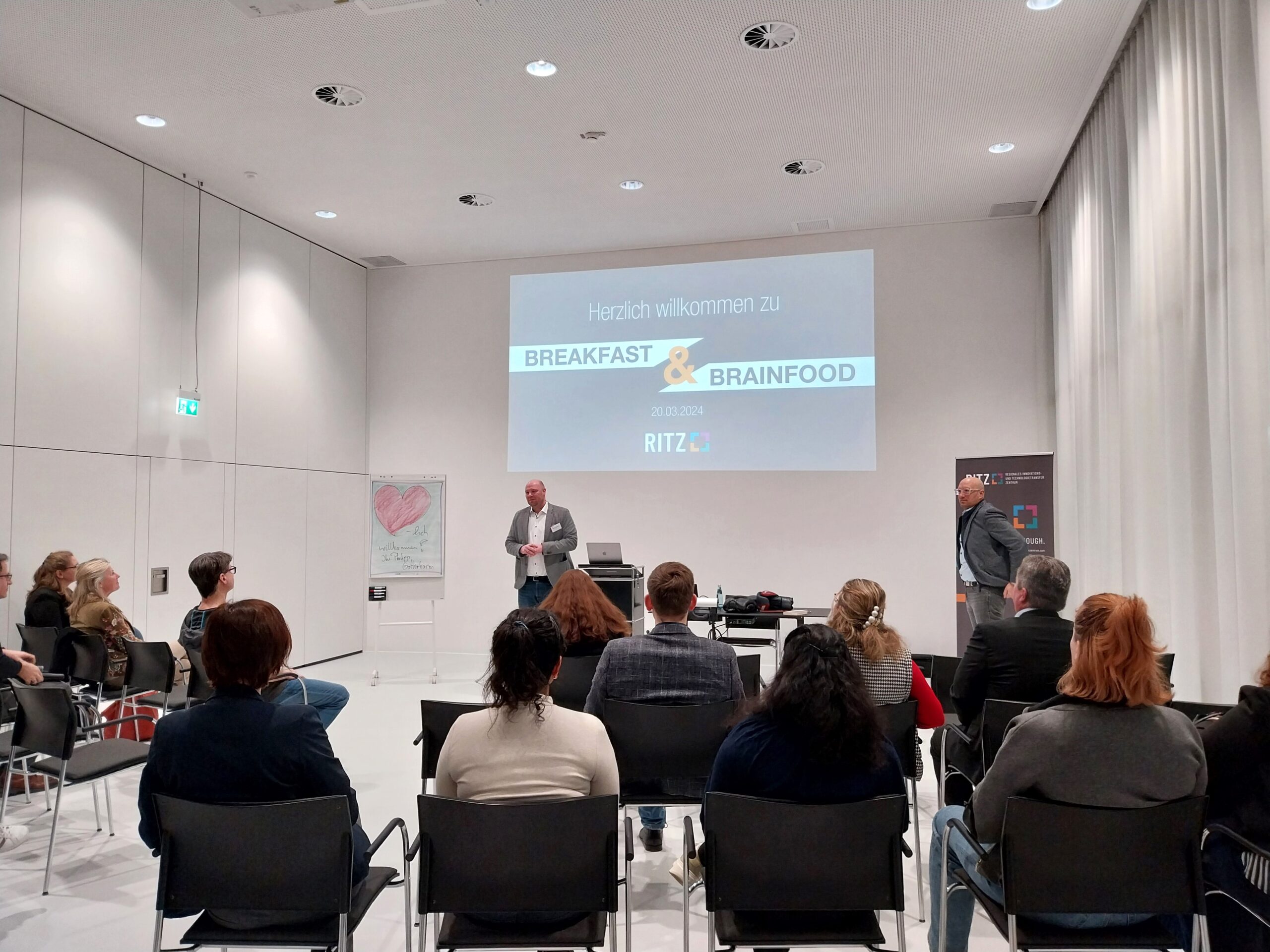 Begrüßung bei der Veranstaltung „Breakfast & Brainfood“ im RITZ, der Geschäftsführer spricht vor sitzendem Publikum, Präsentation im Hintergrund.