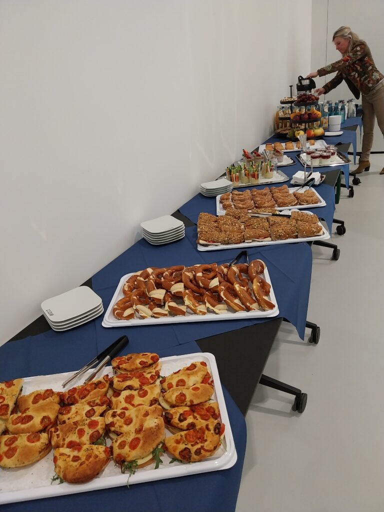 Blick auf das Frühstücksbuffet bei der Veranstaltung ‚Breakfast & Brainfood‘ im RITZ. Auf mehreren Platten liegen belegte Brötchen, Laugenstangen und Tomaten-Focaccia. Im Hintergrund bedient sich eine Frau an der Getränke-Station mit Kaffee.