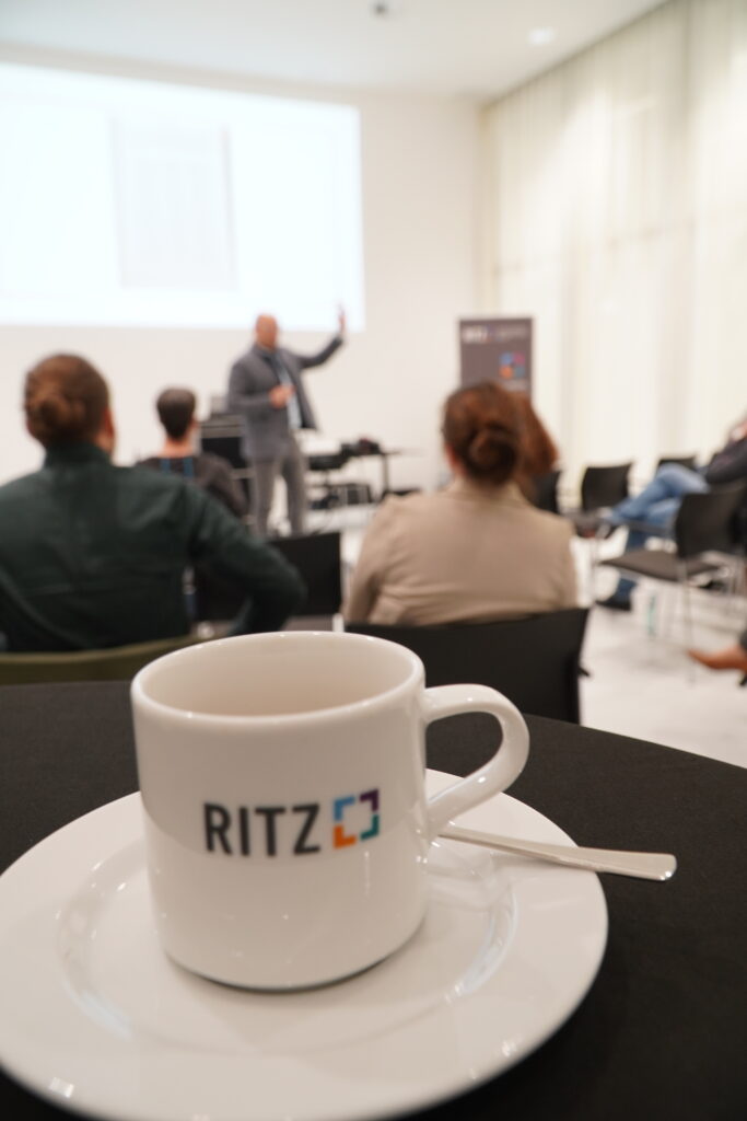 Im Vordergrund eine weiße Kaffeetasse mit RITZ-Logo, im Hintergrund ein unscharfer Vortrag während der Veranstaltung ‚Breakfast & Brainfood‘ im RITZ Friedrichshafen.