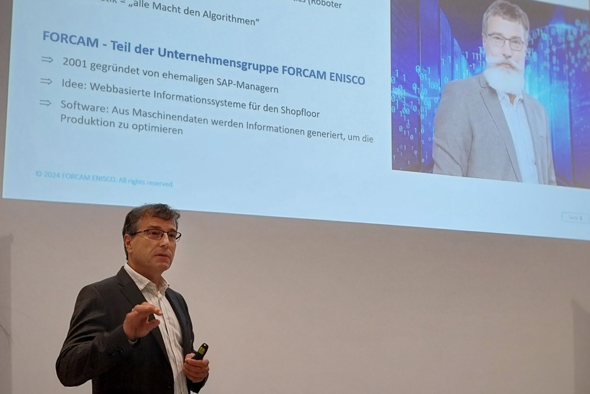 Dr. Alexander Schließmann beim Vortrag im RITZ. Auf der Präsentationsfolie hinter ihm ist ein Porträtfoto eines Mannes mit digital eingefügtem Vollbart zu sehen, daneben Informationen zur Unternehmensgruppe FORCAM ENISCO. Dr. Schließmann spricht zum Publikum und hält einen Presenter in der Hand.