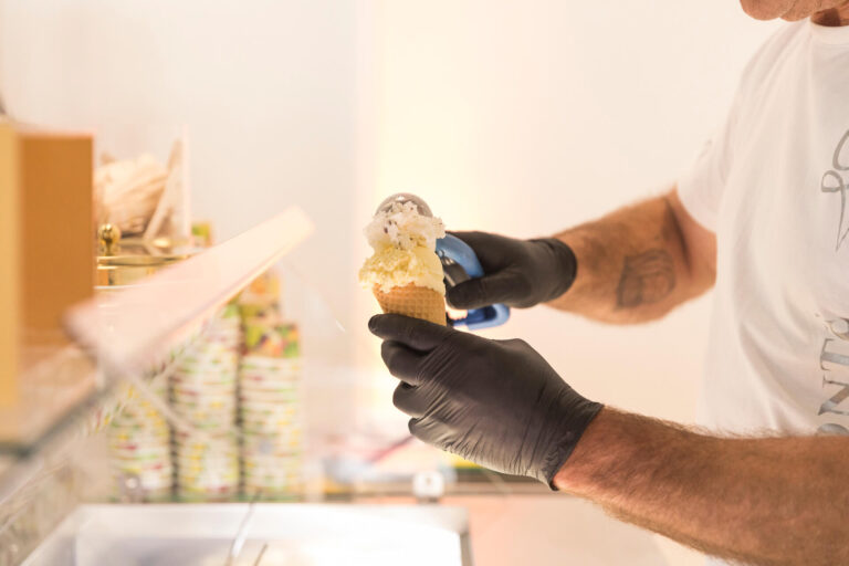 Ein Mitarbeiter der Eisdiele Gelati Conte portioniert zwei Kugeln Eis in eine Waffel. Er trägt schwarze Handschuhe und verwendet eine Eiszange. Im Hintergrund sind bunte Pappbecher gestapelt. Die Szene vermittelt eine sommerliche, genussvolle Atmosphäre beim Event im RITZ.
