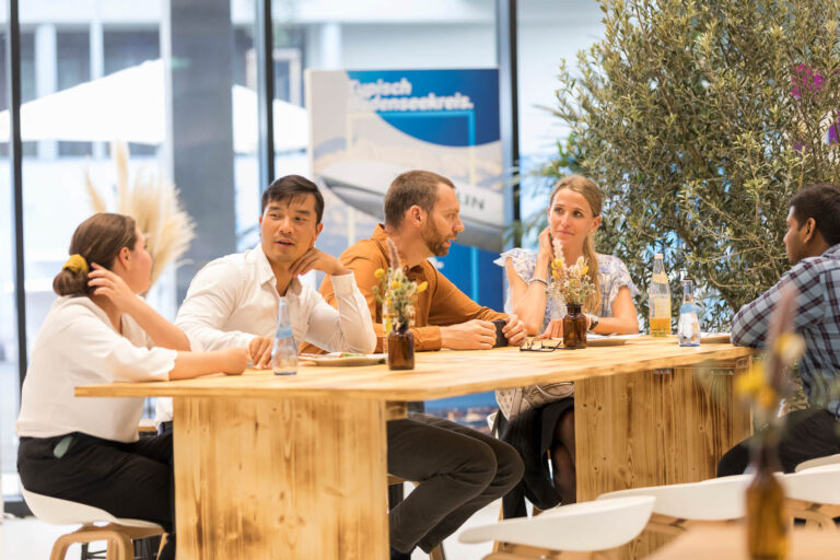 Fünf Personen sitzen an einem langen Holztisch und unterhalten sich lebhaft. Die lockere Atmosphäre wird durch die sommerliche Kleidung, Wasserflaschen, dekorative Blumenvasen und einen Olivenbaum im Hintergrund unterstrichen. Die große Fensterfront sorgt für viel Licht im Raum.