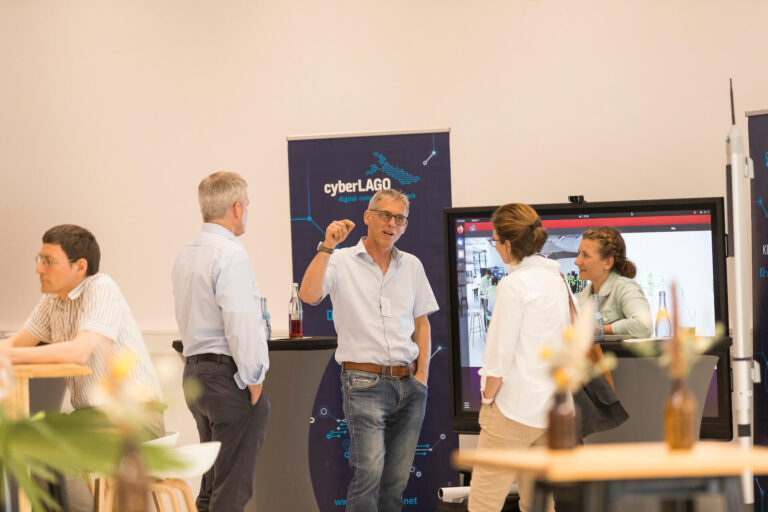 Mehrere Gäste stehen im Ausstellungsbereich vor dem cyberLAGO-Stand und unterhalten sich lebhaft. Im Zentrum des Bildes gestikuliert ein Mann mit Brille und hochgekrempelten Ärmeln. Im Hintergrund sind Roll-up-Banner, ein Monitor und Stehtische zu sehen. Die Szene zeigt informellen Austausch in entspannter Atmosphäre.