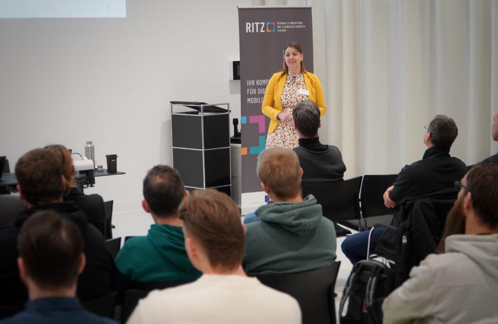 Diplom-Psychologin Julia Kröll beim Vortrag des Breakfast & Brainfood im RITZ. Sie steht vor einem RITZ-Roll-up, trägt ein gelbes Jäckchen über einem geblümten Kleid und spricht zu einem aufmerksam lauschenden Publikum.