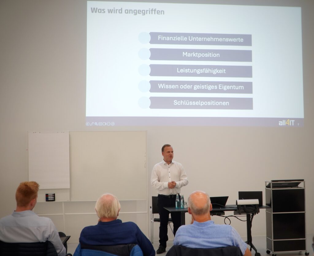 IT-Sicherheitsexperte Matthias Ruf von all4IT beim Vortrag des Breakfast & Brainfood im RITZ. Hinter ihm ist eine Präsentationsfolie zu sehen mit der Frage 'Was wird angegriffen?' und fünf aufgelisteten Unternehmensbereichen. Vor ihm sitzt ein konzentriertes Publikum.