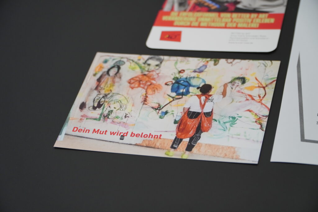 Eine bunte Postkarte zeigt eine Person mit roter Schürze, die vor einer bemalten Wand steht und mit dem Rücken zur Kamera blickt. Auf der Wand sind fantasievolle, abstrakte Zeichnungen und Gesichter zu sehen. Unten links steht in roter Schrift: „Dein Mut wird belohnt“.