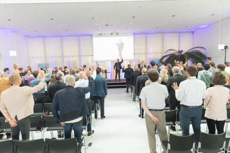 Das Publikum steht im Veranstaltungsraum des RITZ Innovationszentrums und folgt aktiv einer Mitmachübung von Dr. Boris Nikolai Konrad. Alle Teilnehmenden heben synchron die rechte Hand, während der Speaker auf der Bühne die Übung anleitet. Die Stimmung ist lebendig und konzentriert.