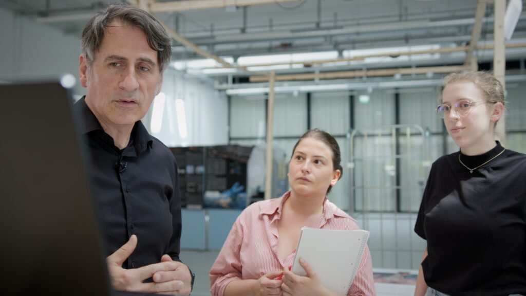Ein Mann und zwei junge Frauen stehen in einem modernen Technikraum. Der Mann erklärt etwas an einem Bildschirm, die Frauen hören aufmerksam zu. Eine von ihnen hält einen Block und einen Stift. Im Hintergrund sind Werkbänke, Kabel und technische Geräte zu sehen.