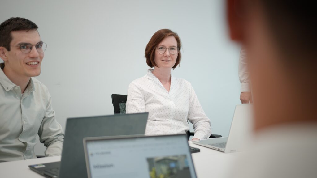 Zwei Personen sitzen an einem Konferenztisch mit Laptops vor sich. Eine Frau in weißer Bluse blickt direkt in die Kamera, ein junger Mann neben ihr lächelt. Im Vordergrund sind unscharf weitere Personen und Laptop-Bildschirme zu sehen.