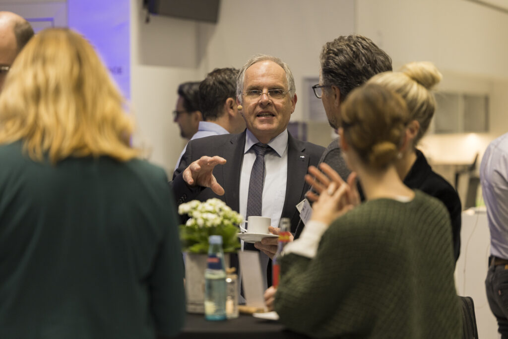 Gesprächsrunde beim Networking: Ein Mann im Anzug mit Headset spricht lebhaft mit mehreren Personen an einem Stehtisch. Auf dem Tisch stehen eine kleine Blume und Getränkeflaschen.