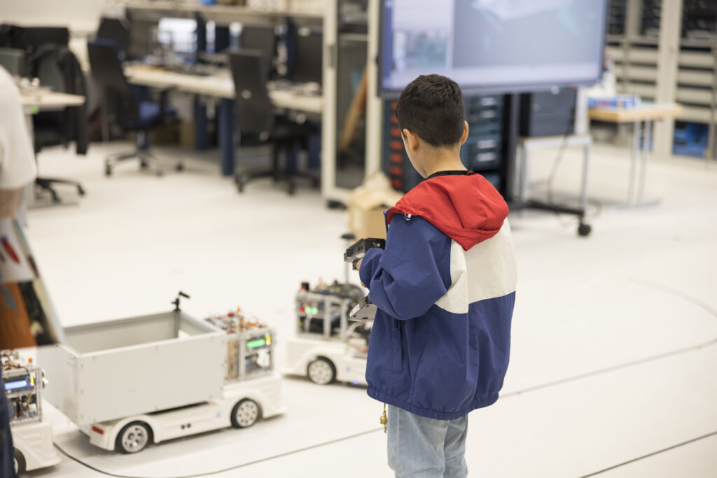 Ein Junge mit blau-weiß-roter Jacke steuert mit einer Fernbedienung ein kleines autonomes Fahrzeug in einem Laborumfeld. Im Hintergrund sind mehrere Arbeitsplätze mit Monitoren sowie ein großer Bildschirm mit technischer Darstellung zu sehen. Die Szene zeigt kindliches Interesse an Technik bei der „Langen Nacht der Technik“.