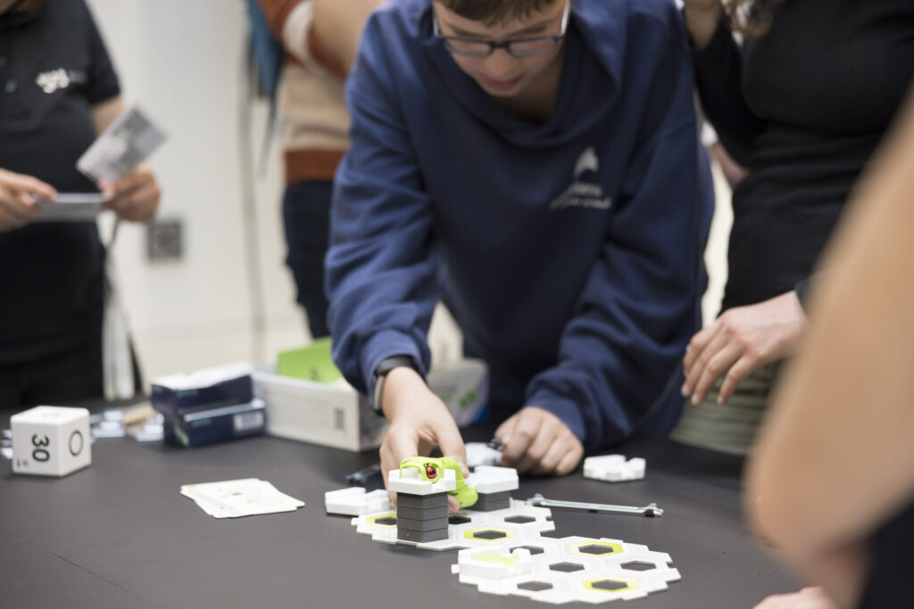 Ein Junge konzentriert sich auf den Aufbau einer Kugelbahn aus hexagonalen Modulen bei der „Langen Nacht der Technik“. Um ihn herum stehen weitere Personen, die das Geschehen beobachten oder mitmachen. Auf dem Tisch liegen Bauteile, Spielkarten und ein Würfel. Die Szene zeigt spielerisches Lernen durch Technik.