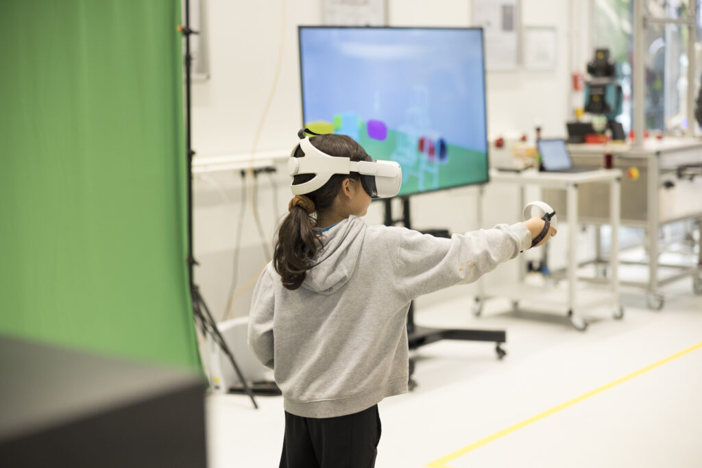 Ein Mädchen steht mit VR-Brille und Controllern in einem Techniklabor. Vor ihr zeigt ein Bildschirm die virtuelle Umgebung, in der sie sich bewegt. Links ist ein Green-Screen zu sehen. Die Szene vermittelt spielerisches Lernen und digitale Interaktion bei der „Langen Nacht der Technik“.