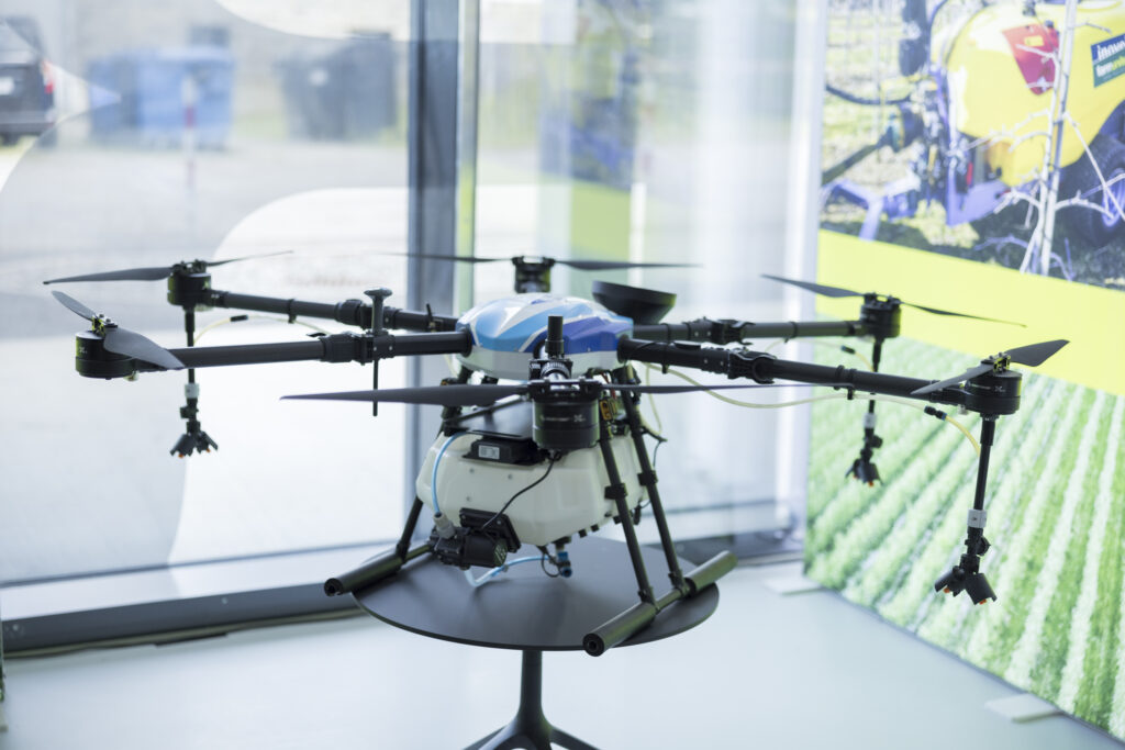 Eine große Drohne mit sechs Rotoren steht auf einem Tisch vor einem Fenster. Im Hintergrund ist ein Banner mit einem landwirtschaftlichen Motiv zu sehen. Die Drohne wirkt professionell und technisch komplex, vermutlich für den Einsatz in der Agrartechnik konzipiert.