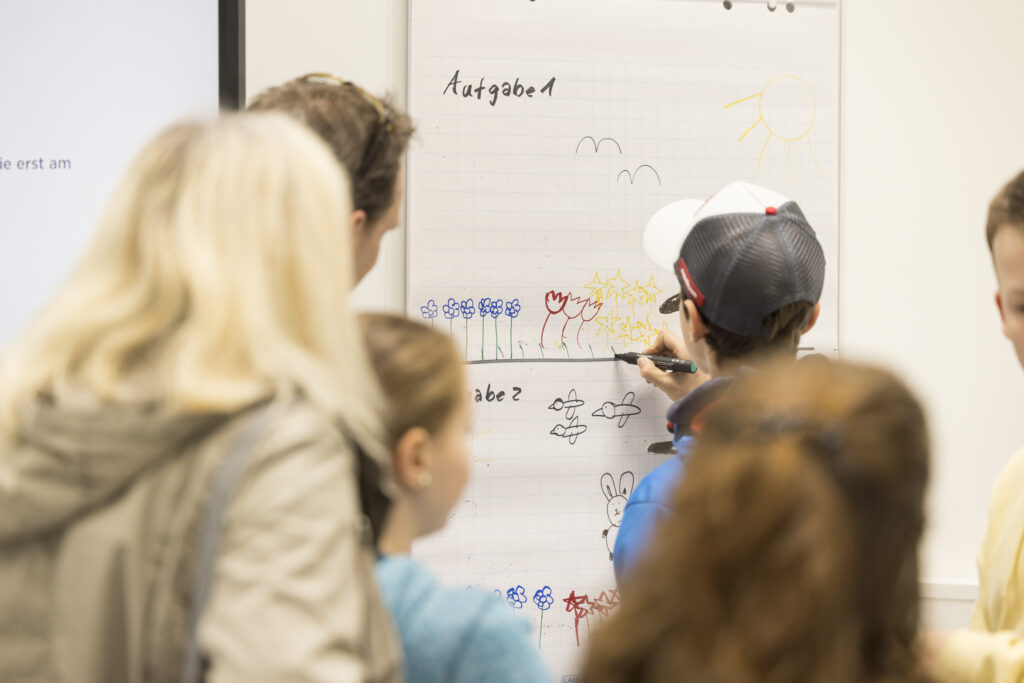 Kinder und Erwachsene stehen vor einem Flipchart, auf dem Aufgaben mit bunten Blumen, Tieren und Symbolen gezeichnet sind. Ein Junge mit Basecap malt konzentriert mit einem schwarzen Filzstift. Die Szene wirkt lebendig und kreativ, als Teil eines Mitmach-Angebots bei einer Veranstaltung.