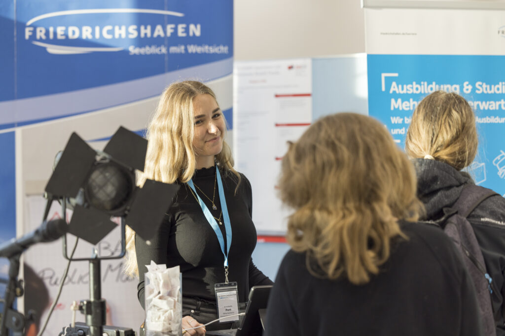 Eine junge Frau mit Namensschild steht am Stand der Stadt Friedrichshafen bei der Langen Nacht der Technik und Innovation (LNTI) und spricht mit zwei Besucherinnen. Im Hintergrund sind Banner zu Ausbildung und Studium sichtbar. Die Szene vermittelt eine offene und einladende Messeatmosphäre.