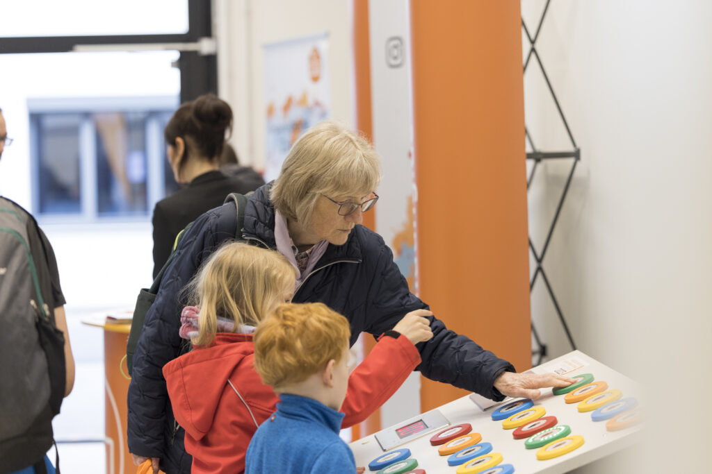 Eine ältere Frau und zwei Kinder interagieren bei der Langen Nacht der Technik und Innovation (LNTI) an einem farbenfrohen Mitmach-Stand. Sie drücken auf große, bunte Knöpfe. Der Stand ist orange gestaltet. Im Hintergrund sind weitere Besucher zu sehen. Die Szene wirkt einladend und familienfreundlich.