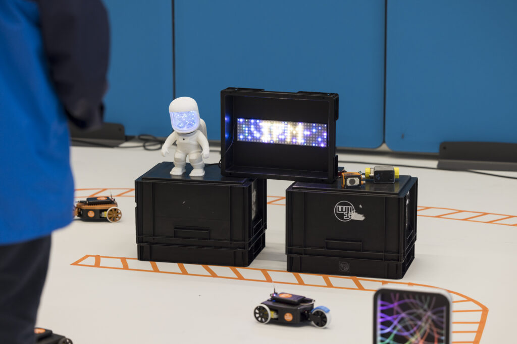 Bei der Langen Nacht der Technik und Innovation (LNTI) steht ein kleiner, beleuchteter Astronaut auf zwei schwarzen Kisten neben einem LED-Display. Um ihn herum fahren Miniroboter auf einer markierten Bahn. Die Szene zeigt ein spielerisches Technik-Setup – ein Mitmachangebot für Kinder rund um Robotik oder Programmierung.