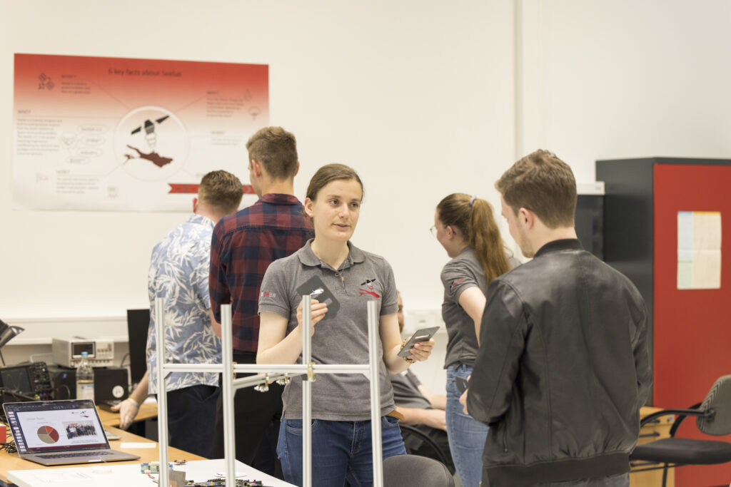 Bei der Langen Nacht der Technik und Innovation (LNTI) erklärt eine junge Frau im grauen Poloshirt mit Logo des studentischen Satellitenprojekts SeeSat einem Besucher zwei Bauteile. Im Hintergrund arbeiten Teammitglieder an Laptops, auf einem Poster sind zentrale Fakten zum Projekt dargestellt. Die Szene vermittelt praxisnahes Technikverständnis und engagierten Wissensaustausch.