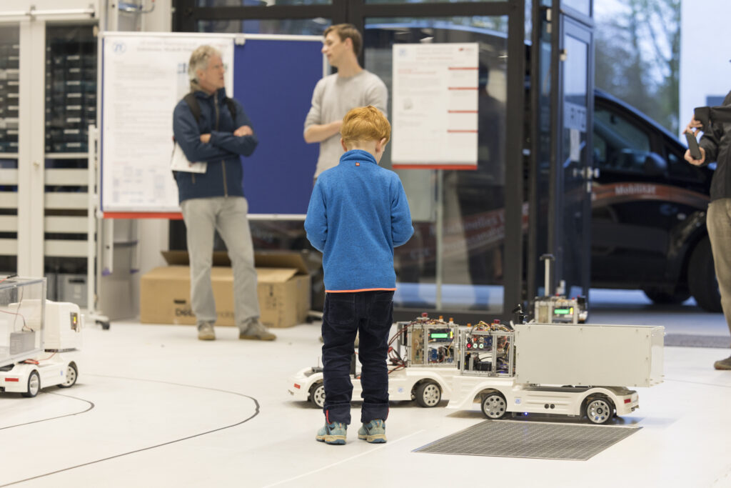 Kind steuert bei der Langen Nacht der Technik und Innovation (LNTI) im RITZ ein autonom fahrendes Modellfahrzeug. Das Fahrzeug ist mit Sensoren und Elektronik ausgestattet. Im Hintergrund informieren sich Erwachsene an Posterwänden über das Forschungsprojekt.