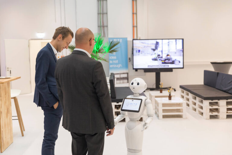 Der humanoide Roboter Pepper begrüßt zwei männliche Gäste im Ausstellungsbereich des RITZ Innovationszentrums. Auf seinem Bildschirm ist das RITZ-Logo sichtbar. Im Hintergrund stehen Palettenmöbel, Monitore mit Technikvideos und ein Banner mit dem Text „Sometimes an idea is not enough“.