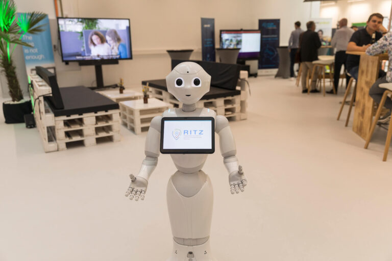 Der humanoide Roboter Pepper steht im Mittelpunkt des Ausstellungsbereichs im RITZ Innovationszentrum. Auf seinem Bildschirm ist das RITZ-Logo zu sehen. Im Hintergrund sind Besuchergruppen, Messestände, Monitore und Lounge-Möbel aus Paletten erkennbar. Die Szene wirkt modern und einladend.
