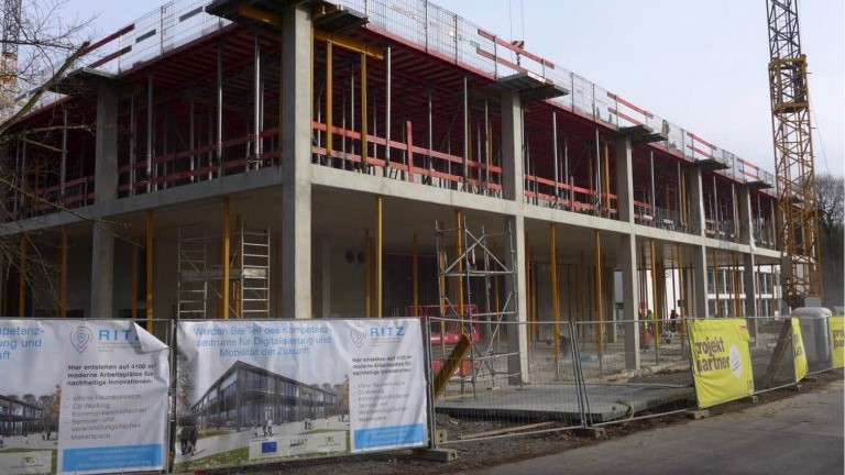 Rohbau des RITZ Innovationszentrums: Zweigeschossiger Betonbau mit Gerüsten, Bauzäunen und Informationsbannern an der Vorderseite; Baukran am rechten Bildrand.