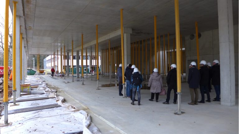 Gruppe von Personen mit Schutzhelmen besichtigt den Rohbau des RITZ Innovationszentrums; Sicht auf betonierte Decke mit gelben Stützen und offene Baufläche.