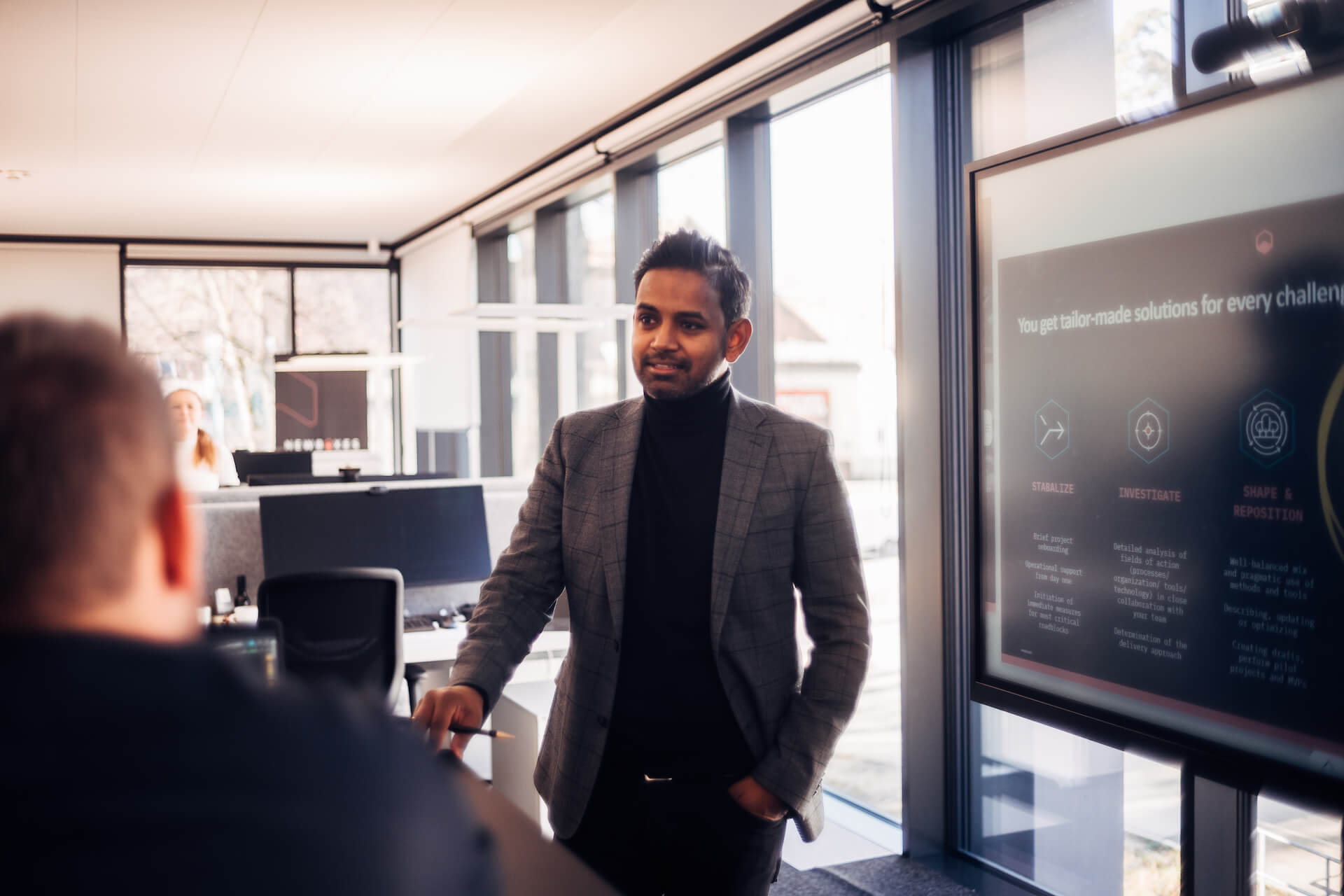 Suwi Murugathas von newboxes steht im Business-Outfit vor einem Bildschirm und erklärt einem Kollegen eine Präsentation – moderne Büroumgebung mit großen Fenstern und hellen Arbeitsplätzen im Hintergrund.