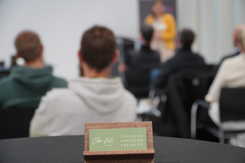 Im Vordergrund liegt eine grüne Visitenkarte mit der Aufschrift ‚Julia Kröll – Coaching, Beratung, Training‘ auf einem Tisch. Im Hintergrund unscharf: Teilnehmer und Vortragende beim Breakfast & Brainfood im RITZ.