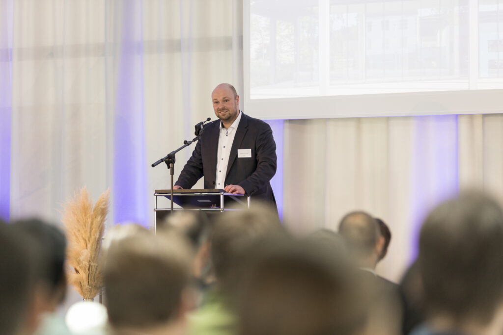 Geschäftsführer Marian Duram spricht beim anderthalbjährigen Jubiläum des RITZ Innovationszentrum an einem Rednerpult mit Mikrofon vor Publikum. Im Hintergrund eine Leinwand und lichtdurchflutete Vorhänge mit dezentem Lila-Lichtakzent.