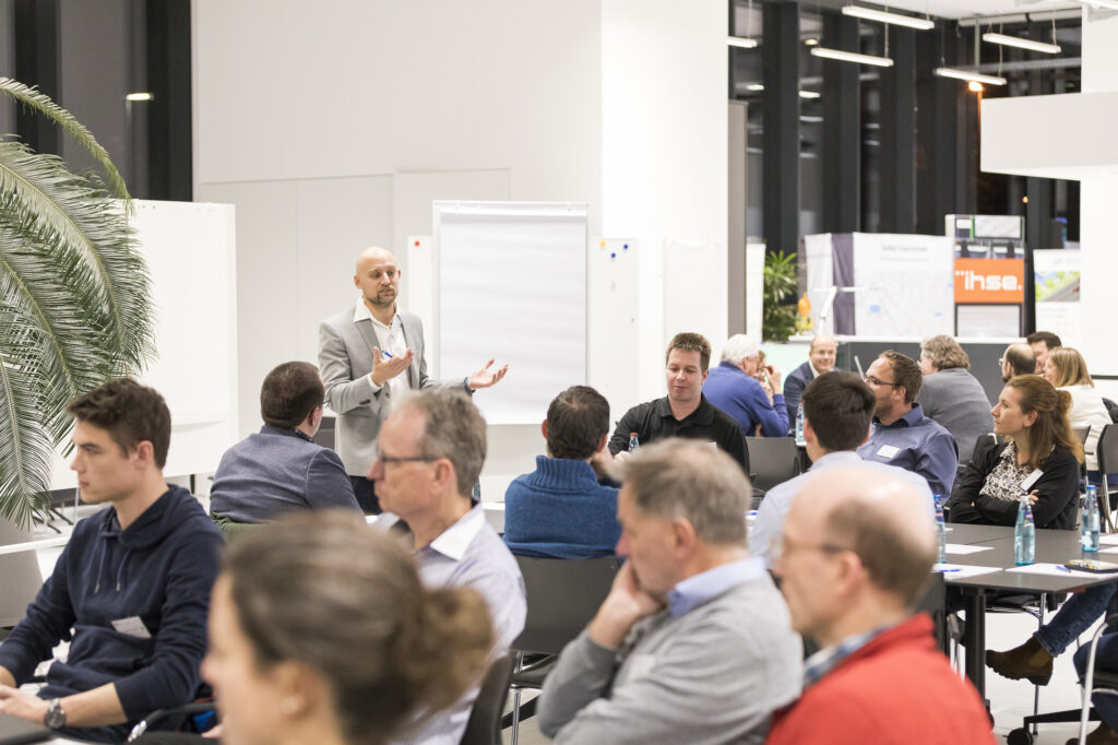 Diskussionsrunde im RITZ Friedrichshafen: Markus Beller von doubleSlash im grauen Sakko spricht vor Teilnehmenden, die an Gruppentischen sitzen. Im Hintergrund sind Präsentationsstände und Whiteboards zu sehen.